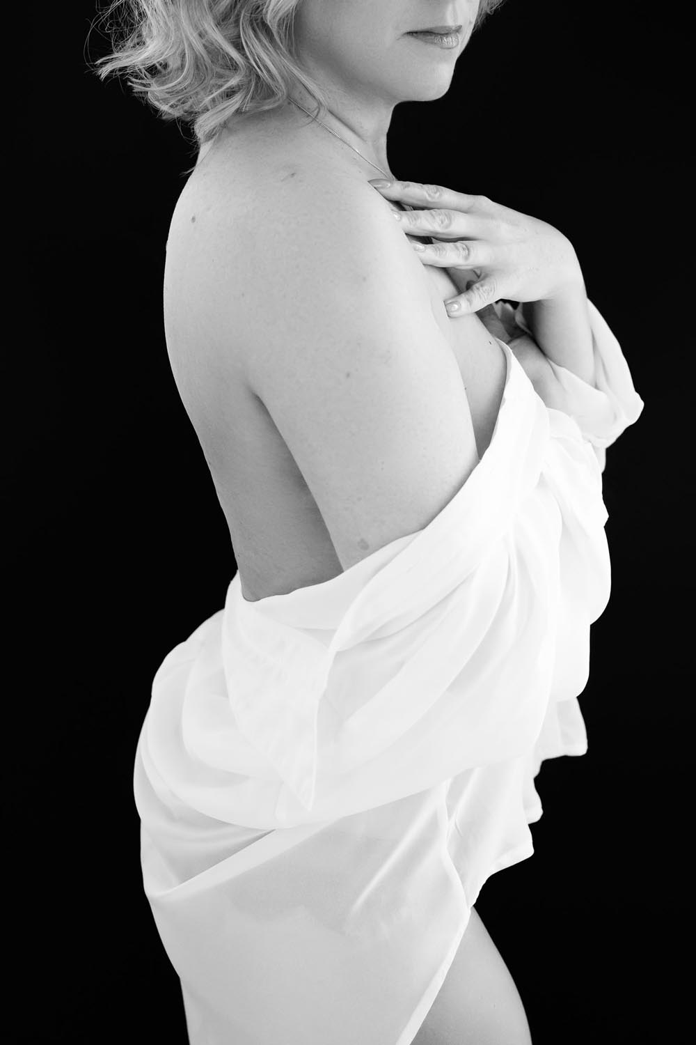 Black and white photo of a woman in profile, draped in a white translucent fabric, with one hand touching her chest and short wavy hair against a black background.