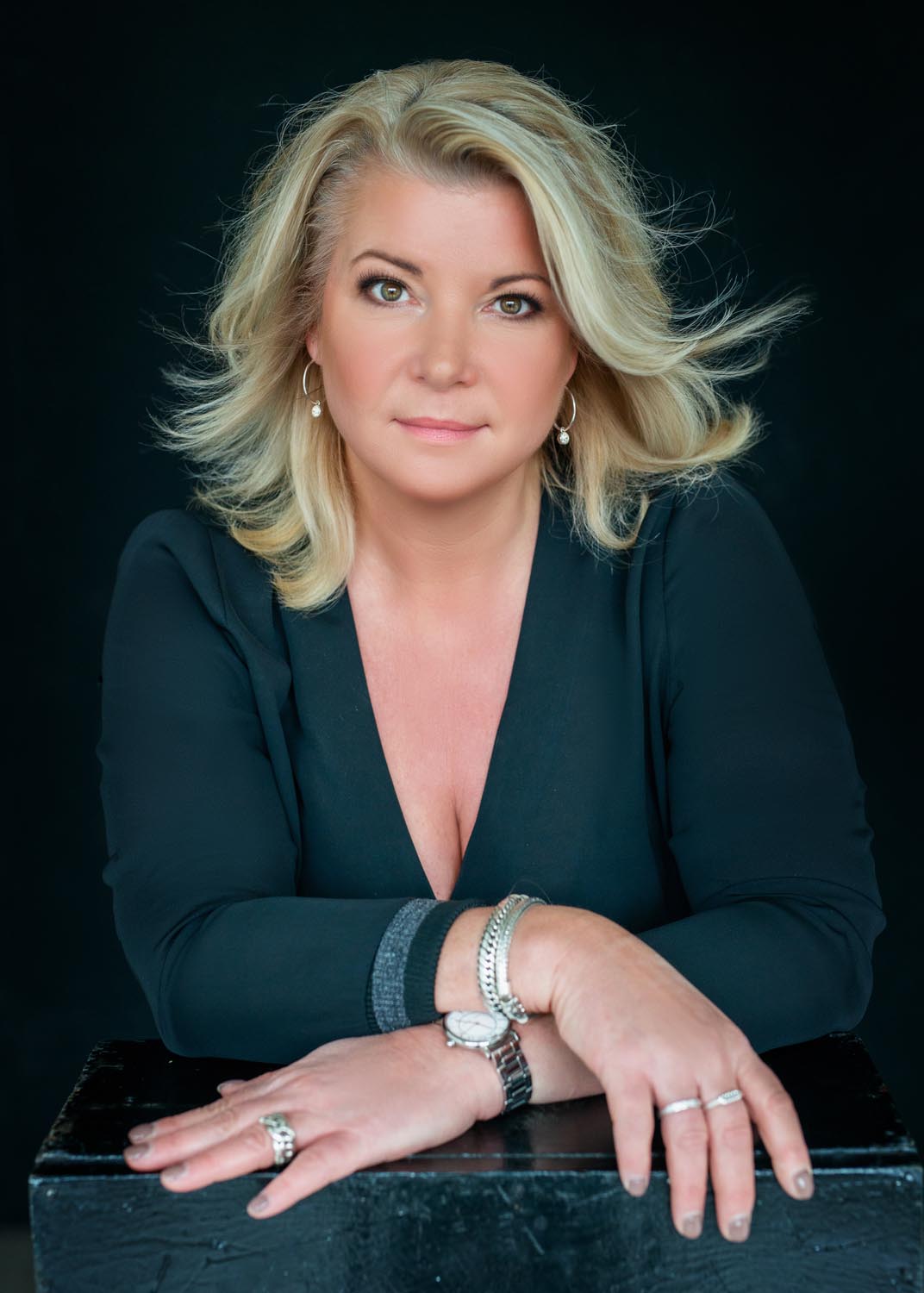 Blonde woman with shoulder-length hair wearing a dark V-neck top, resting her arms on a black surface and looking directly at the camera against a black background.