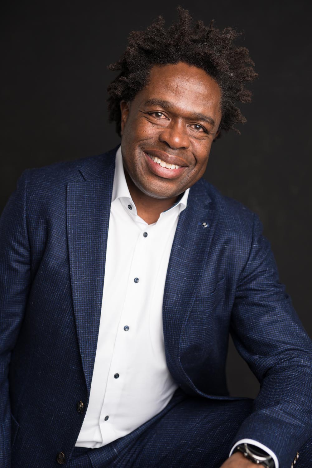 Smiling man with dark curly hair wearing a navy blue patterned suit and white dress shirt, posing against a black background.