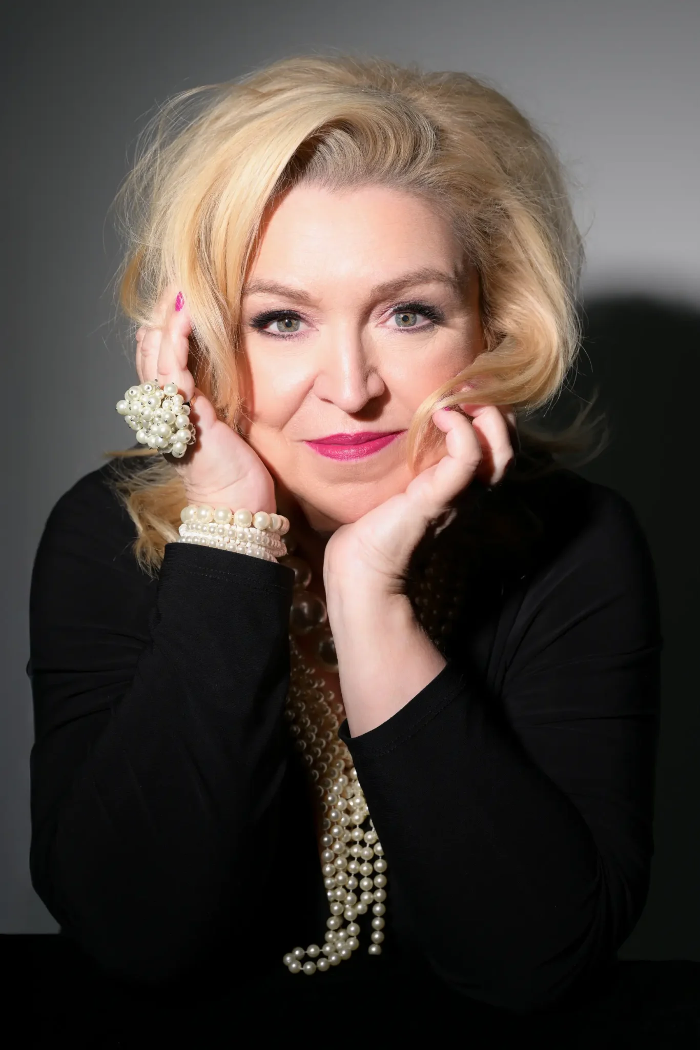 A woman with blonde hair and light green eyes wearing bright pink lipstick, posing with hands near her face. She is dressed in black and adorned with multiple pearl necklaces, bracelets, and a large pearl ring, against a plain gray background.
