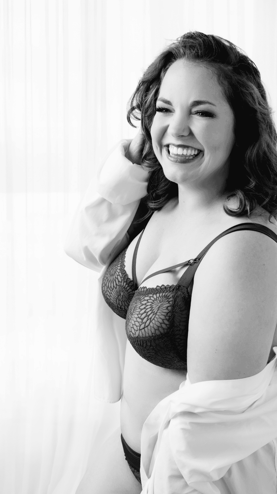 Smiling woman with curly hair wearing a black lace bra and underwear, partially covered by a white shirt, standing in front of a bright window with sheer curtains.