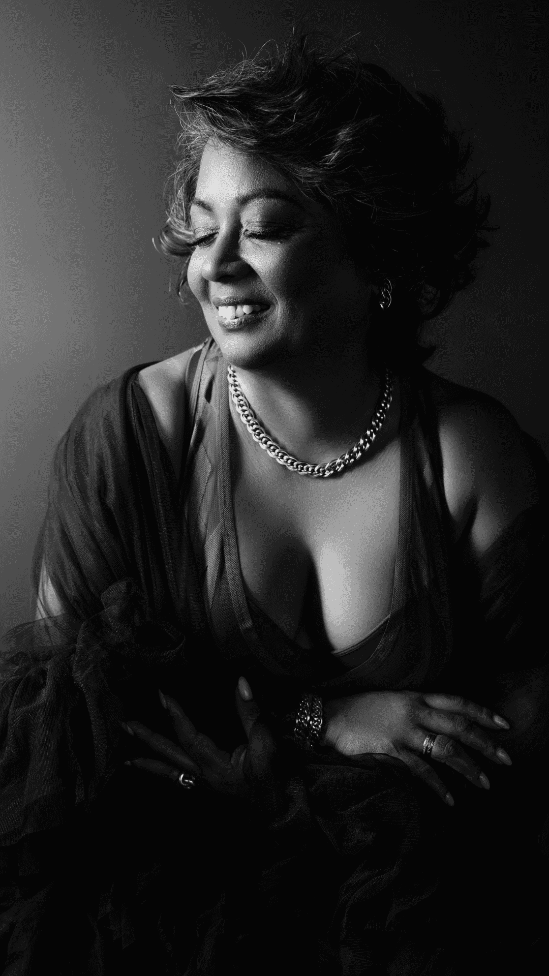 Black and white portrait of a smiling woman with short wavy hair looking to the side, wearing a chain necklace, earrings, a low-cut top, and a sheer shawl draped over her shoulders.