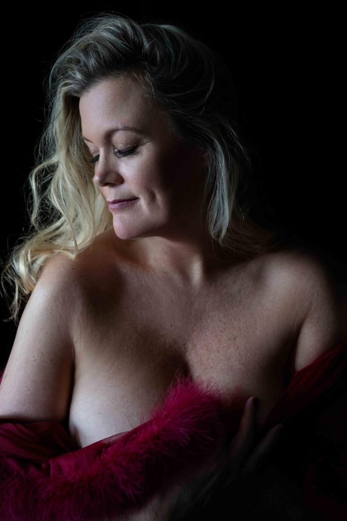 Portrait of a woman with long blonde hair looking down and smiling softly, partially draped in a red feathered garment, against a black background.