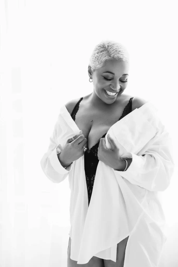 Smiling plus-size woman with short blonde hair wearing a white shirt over black lace lingerie, holding the shirt open and looking down.