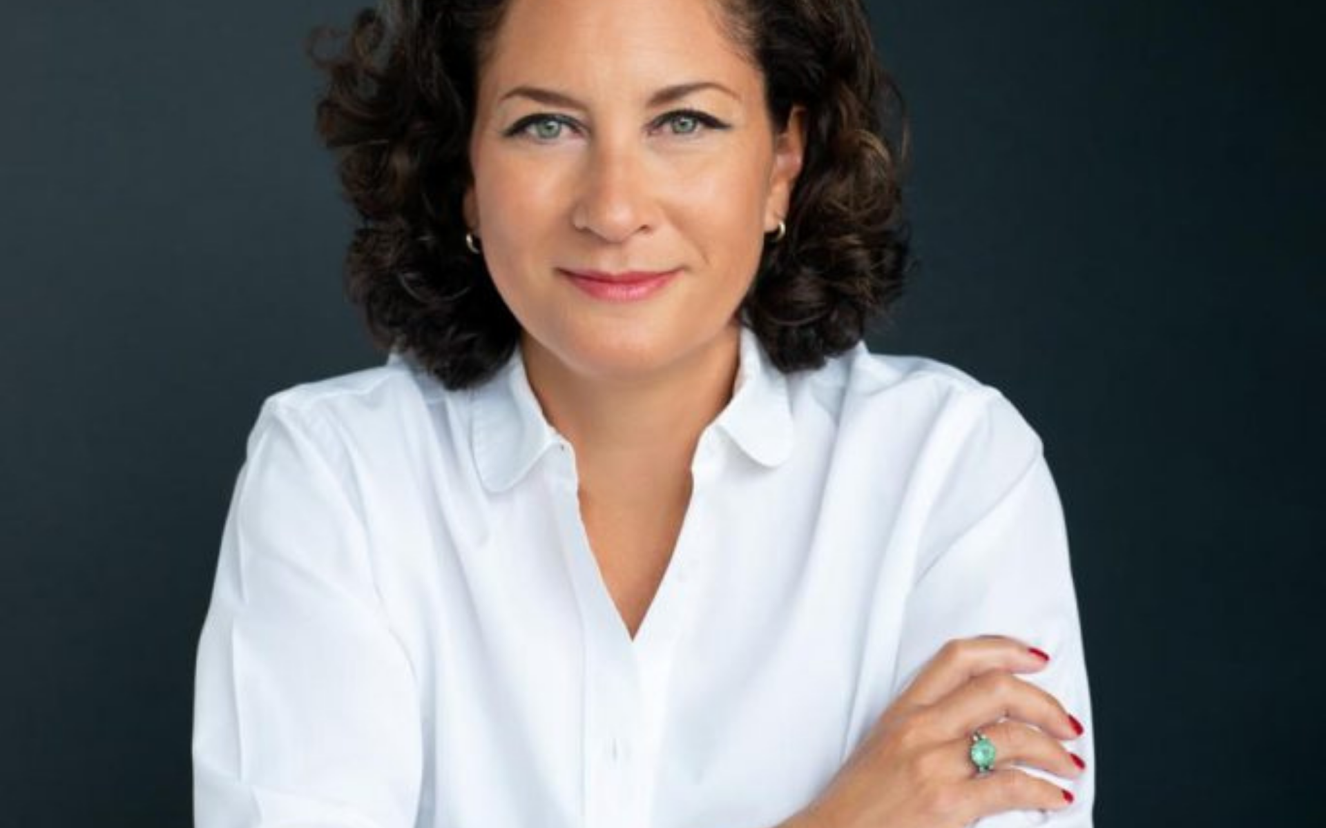 Portrait of a woman with curly dark hair and green eyes wearing a white button-up shirt, small hoop earrings, and a ring with a green stone on her finger, smiling slightly against a dark background.