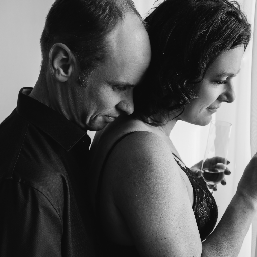 Black and white photo of a man and woman standing close, the man resting his head on the woman's shoulder while she holds a glass and looks down.