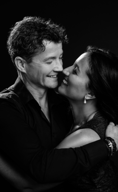 Black-and-white portrait of a smiling couple embracing closely, about to kiss, against a dark background.