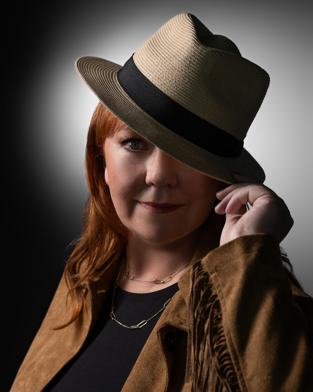 Portrait of a woman with red hair wearing a tan fedora hat tilted over one eye, a brown suede jacket with fringe detailing, a black top, and two gold chain necklaces, posing against a dark gradient background.
