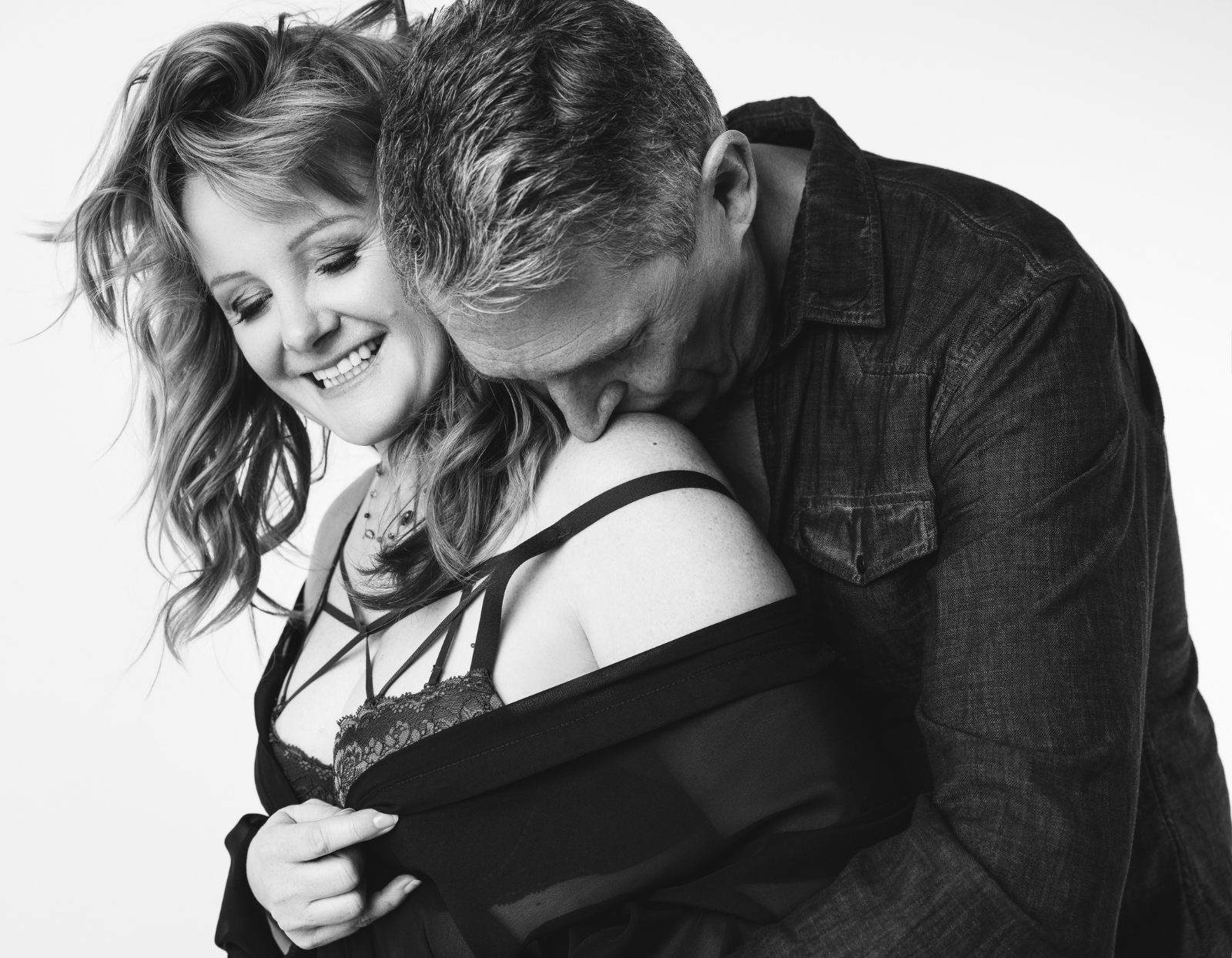 Black and white photo of a smiling woman with loose hair wearing a lace top and man embracing her from behind and kissing her shoulder.