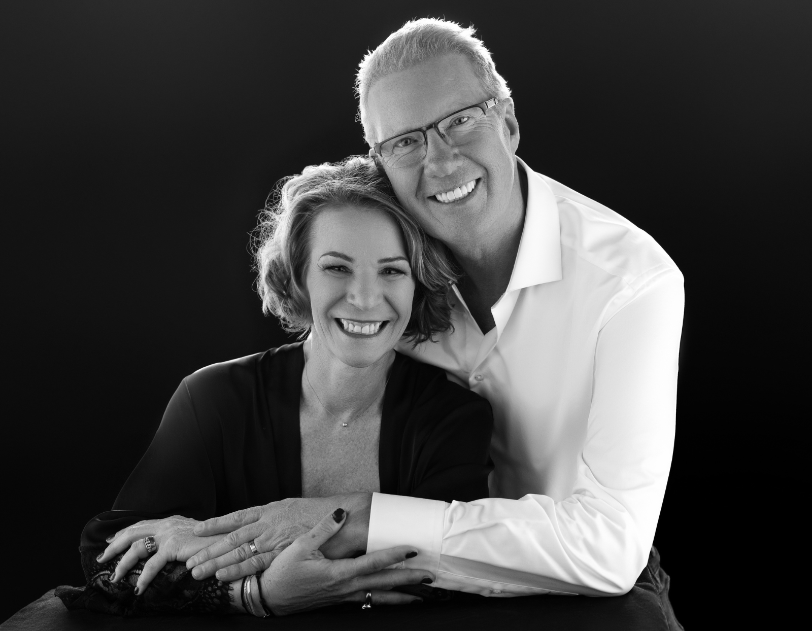Black and white portrait of a smiling middle-aged couple embracing each other against a dark background.