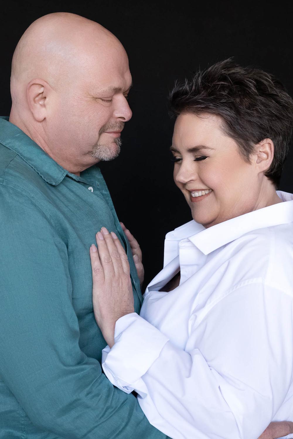 Middle-aged couple facing each other closely, smiling warmly, with the woman’s hands resting gently on the man's chest; the man wears a teal shirt and the woman a white blouse, against a black background.