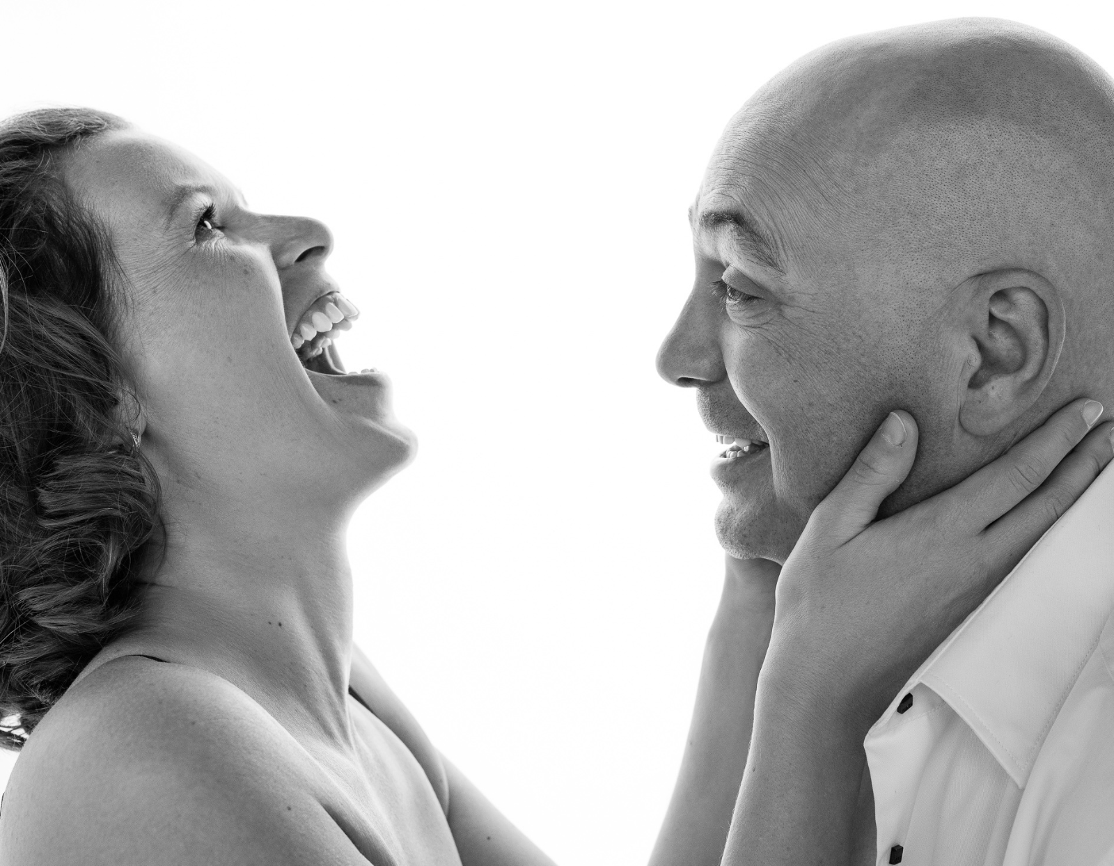 Black and white close-up of a woman and a man facing each other; the woman is laughing joyfully while holding the man's face, and the man smiles back at her.