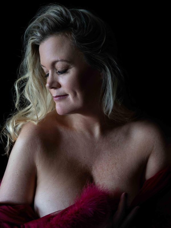 Woman with long blonde hair looking down, partially covered with a red furry fabric against a black background.