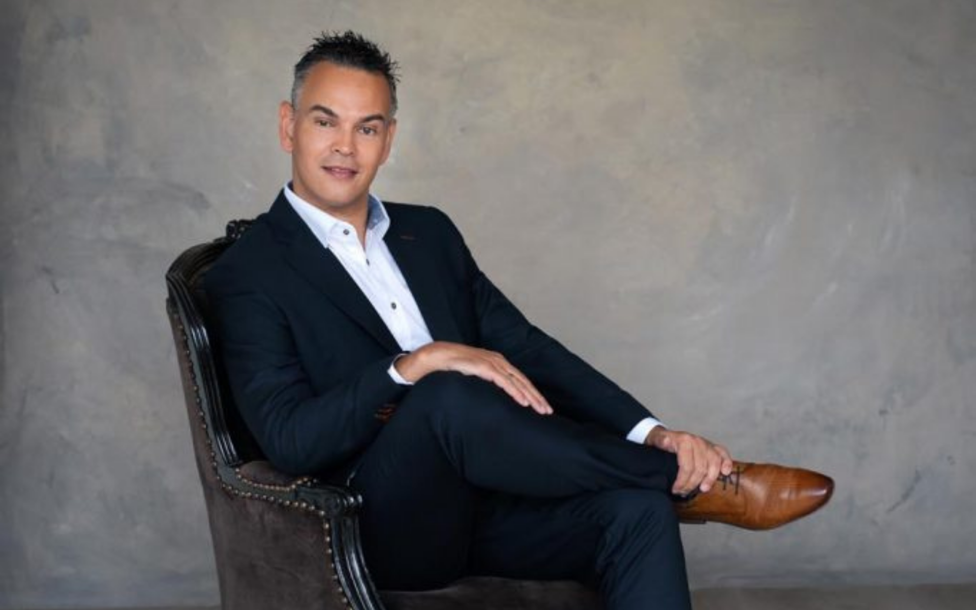 Man in a dark suit and white shirt sitting cross-legged in an armchair against a gray textured wall.