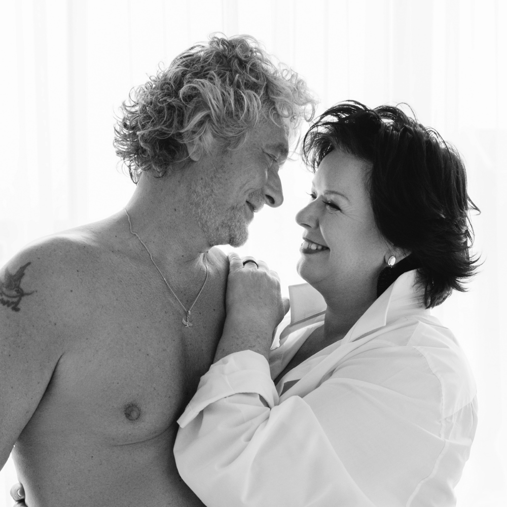 Black and white photo of a smiling man with curly hair and a necklace embracing a woman in a white shirt, both looking into each other's eyes.