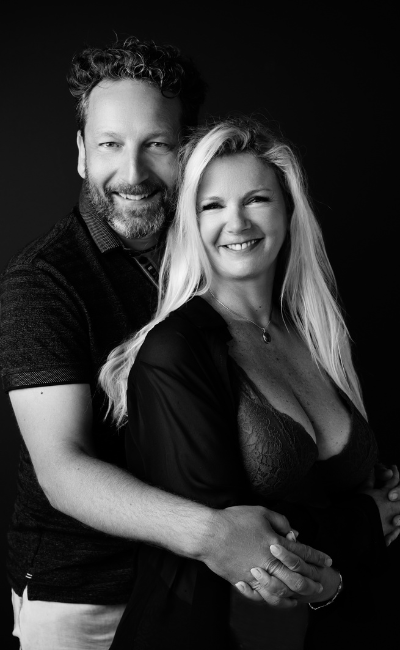 Black and white portrait of a smiling man and woman embracing, with the man standing behind the woman holding her hands.