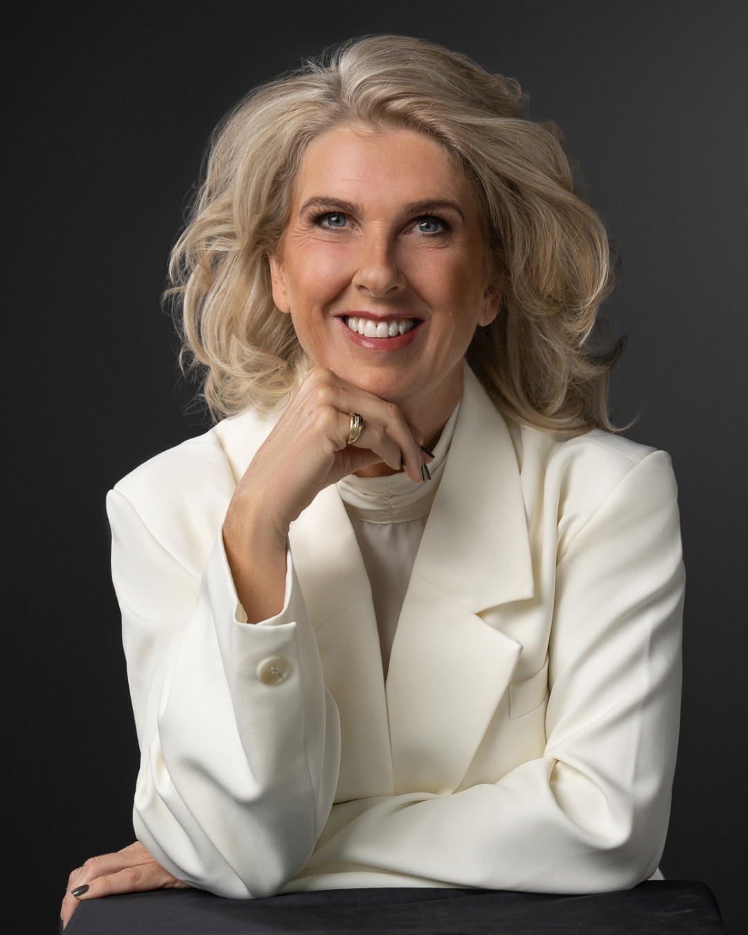 Smiling middle-aged woman with blonde hair wearing a white blazer, resting her chin on her hand against a dark background.