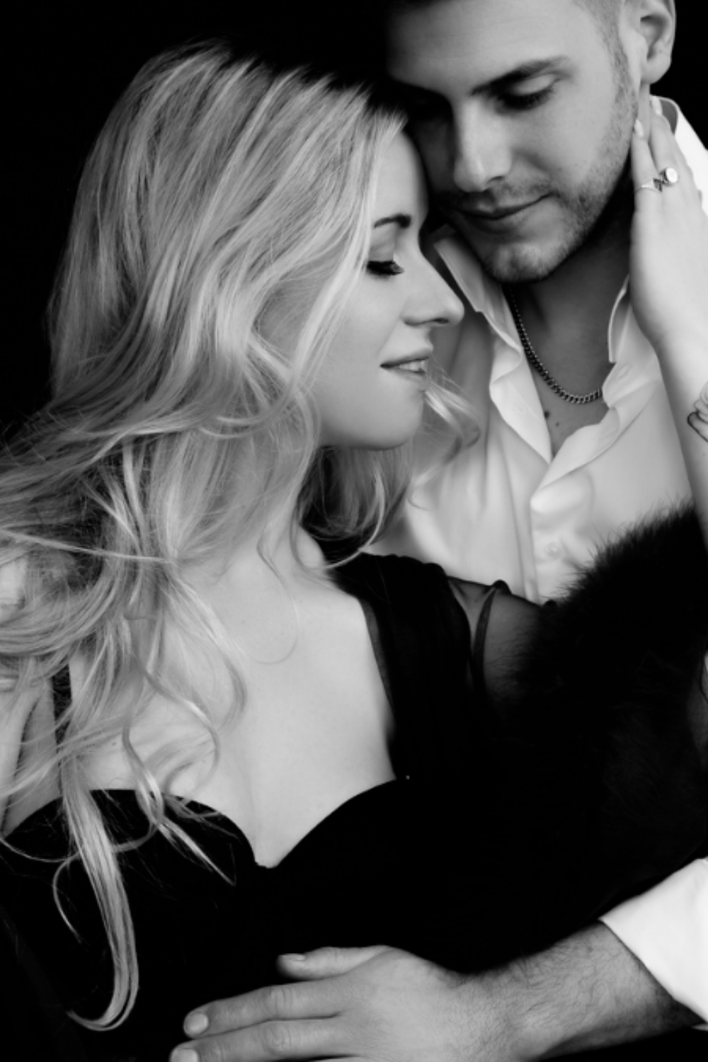 Black and white close-up of a couple embracing intimately, the woman with long blonde hair wearing a black off-shoulder dress and the man in a white shirt with a chain necklace, both with eyes closed and serene expressions.