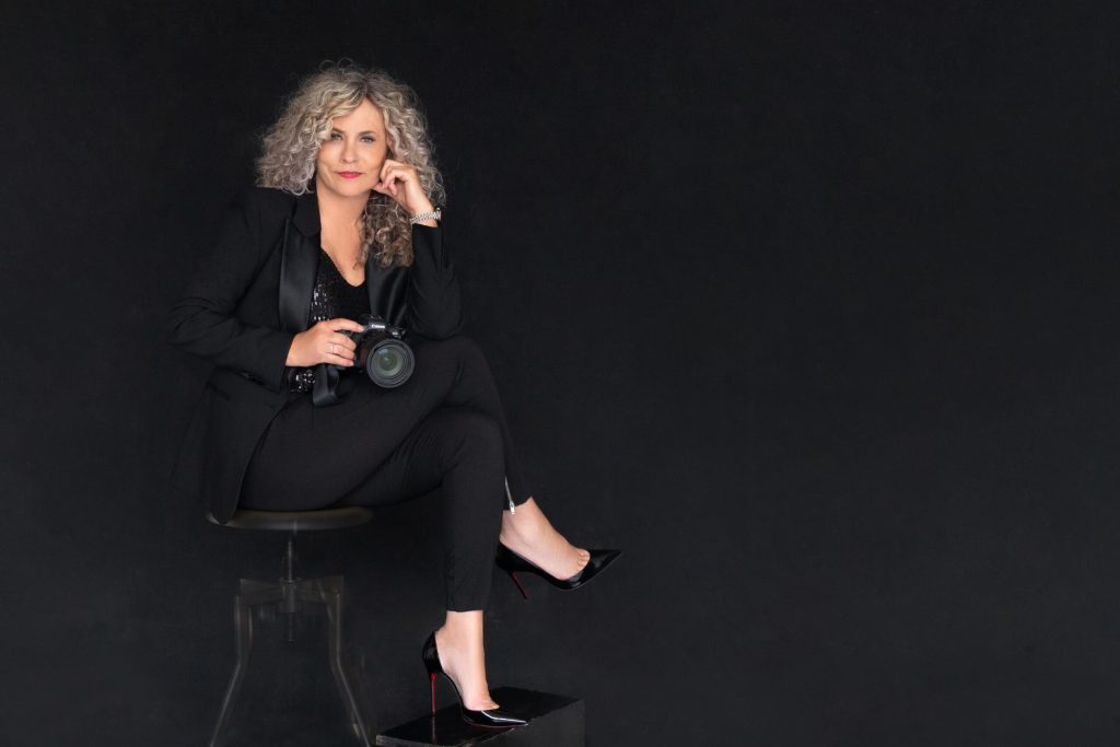 Woman with curly blonde hair wearing a black blazer and pants sitting on a stool against a black background, holding a camera and resting her head on her hand.