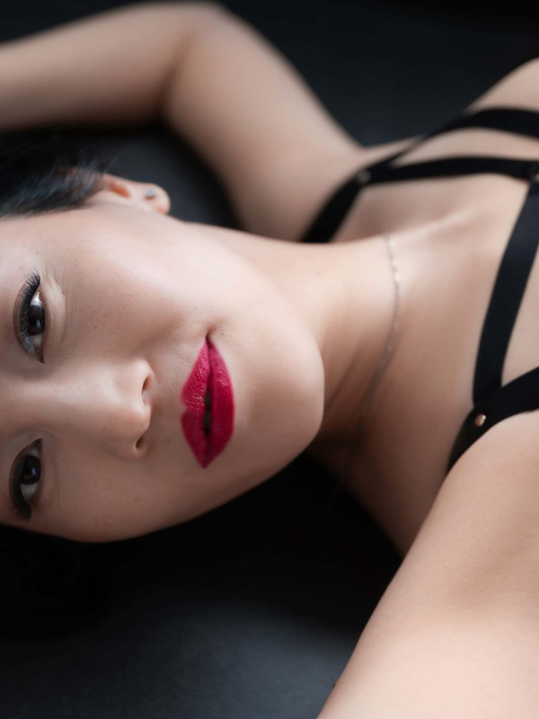 Close-up of a woman lying down with red lipstick, wearing a black strappy top and a thin necklace, her eyes open and gazing ahead.