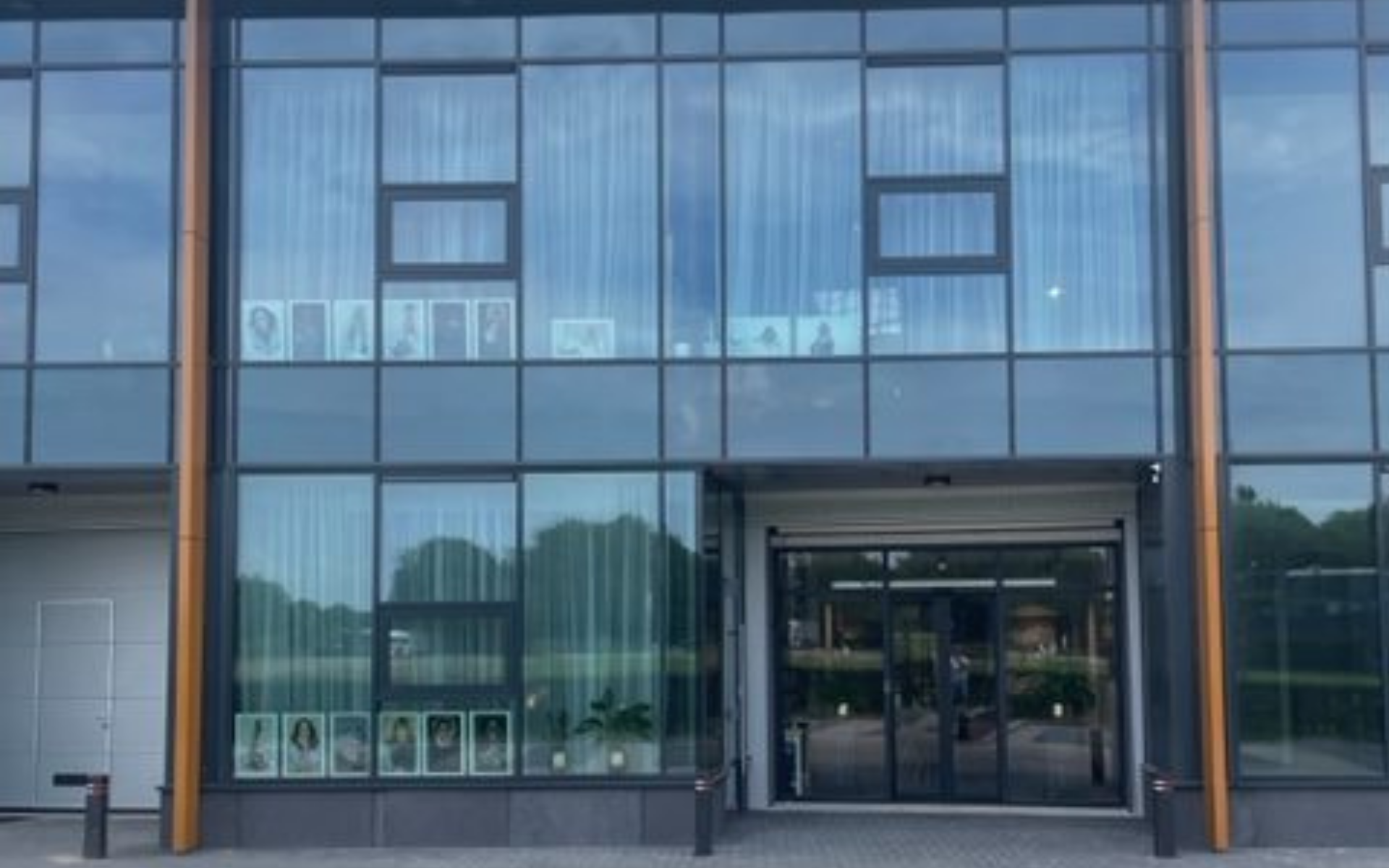 Modern building facade with large glass windows reflecting trees and sky, with some framed photos displayed on the lower and upper window sections, and a glass entrance door in the center.