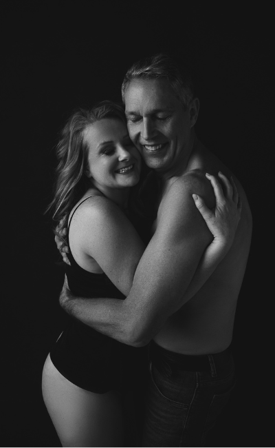 Black and white photo of a smiling couple with eyes closed hugging each other in a warm embrace against a dark background; the woman wears a sleeveless top and the man is shirtless.