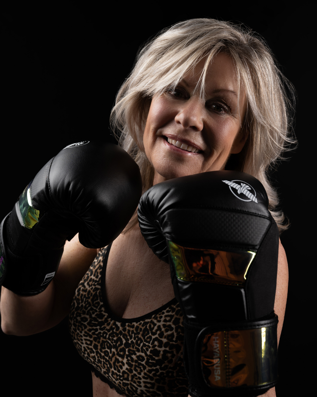 Smiling mature woman with blonde hair wearing black boxing gloves and a leopard-print sports bra, posing in a boxing stance against a black background.