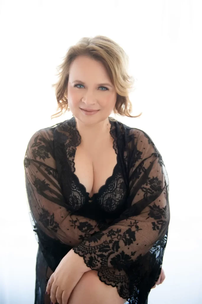 Smiling woman with light brown hair wearing a black lace robe sitting with arms crossed over her knees against a bright white background.