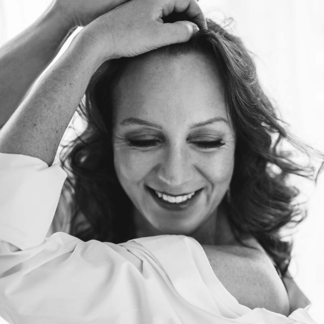 Black and white close-up photo of a smiling woman with closed eyes, gently touching her hair with one hand while wearing an off-shoulder white shirt.