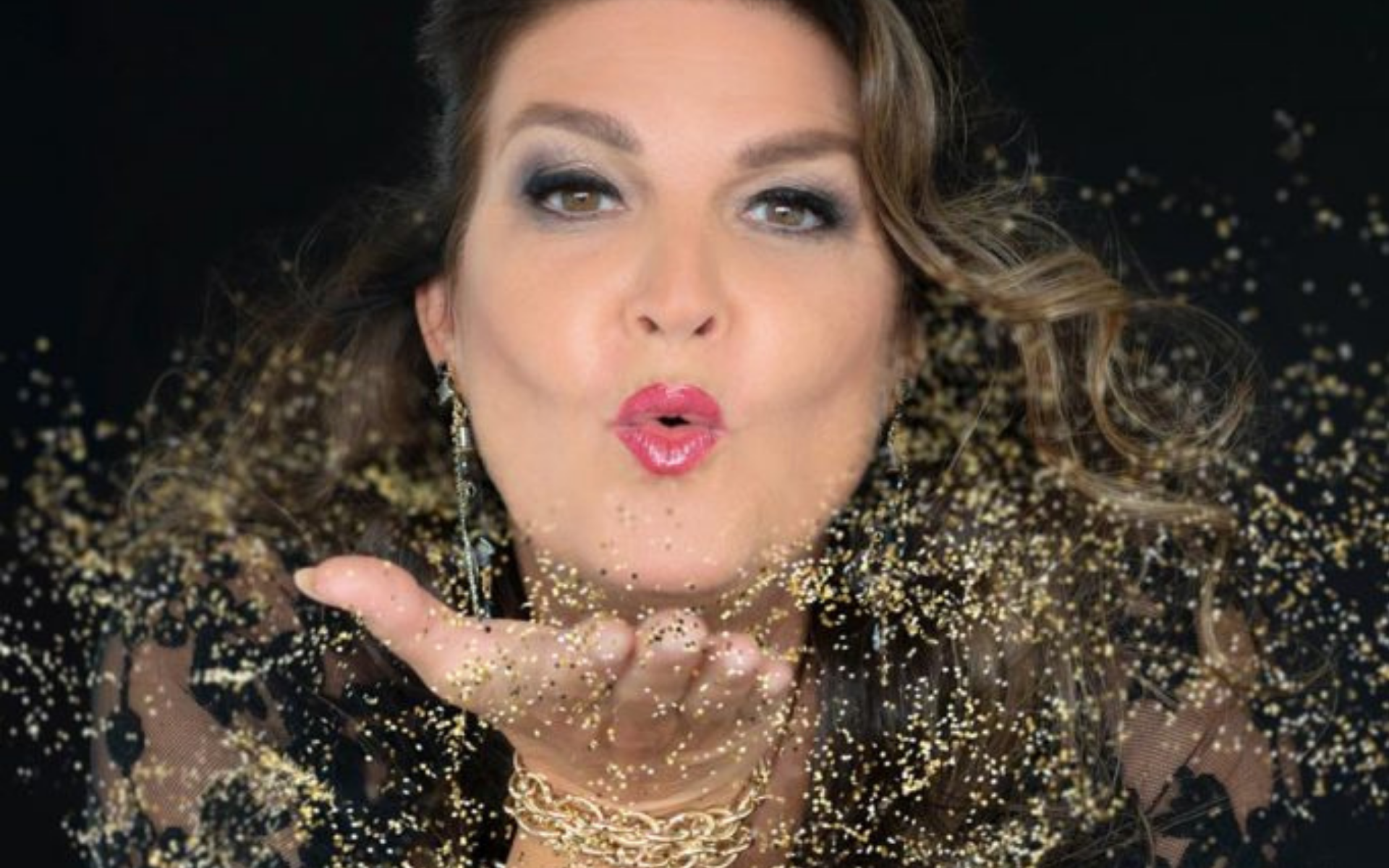 Woman with wavy brown hair blowing gold glitter from her hand, wearing dark eye makeup, bright pink lipstick, and gold jewelry against a black background.
