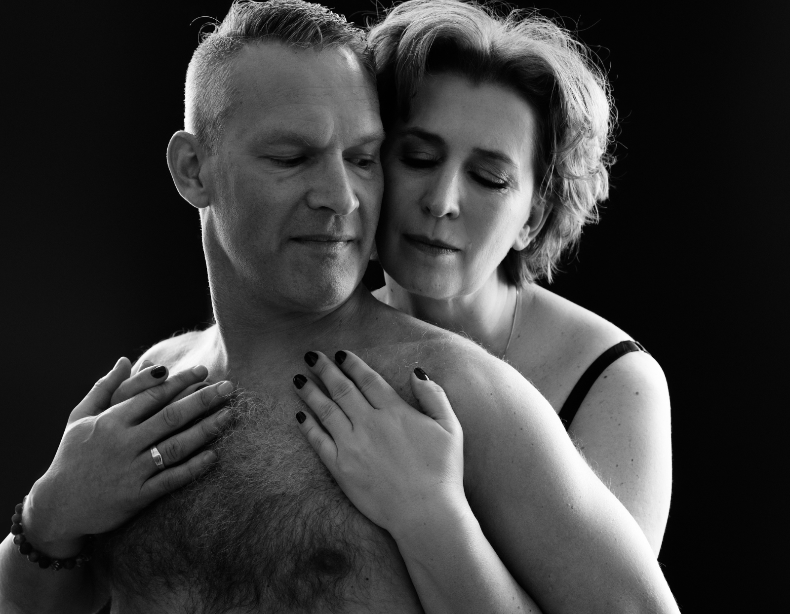 Black and white photo of a woman embracing a shirtless man from behind with both placing their hands on his chest, conveying affection and closeness.