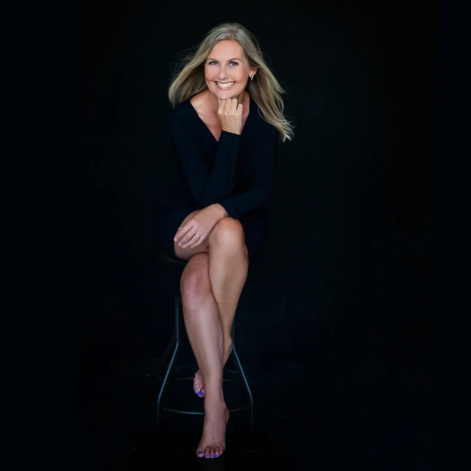 Smiling blonde woman with blue eyes wearing a black dress, sitting cross-legged on a stool against a black background, resting her chin on her hand.