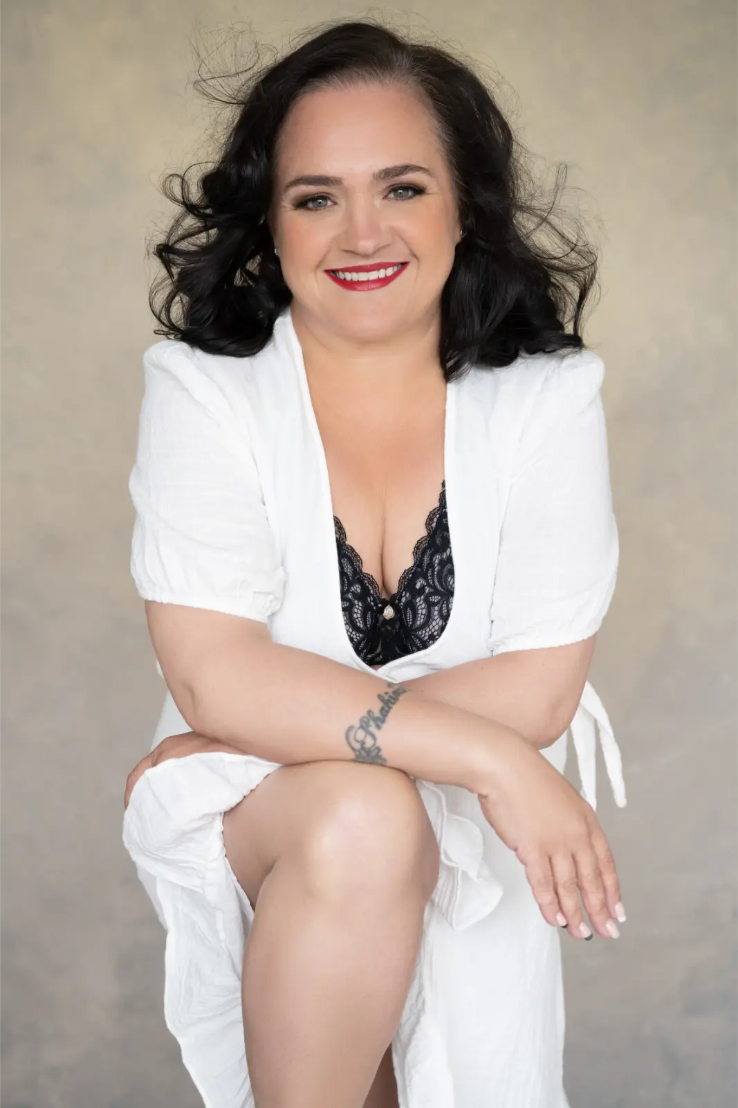 Smiling woman with dark curly hair wearing a white dress and black lace top, sitting with one knee raised and crossing her arms over it, showing a tattoo on her forearm.