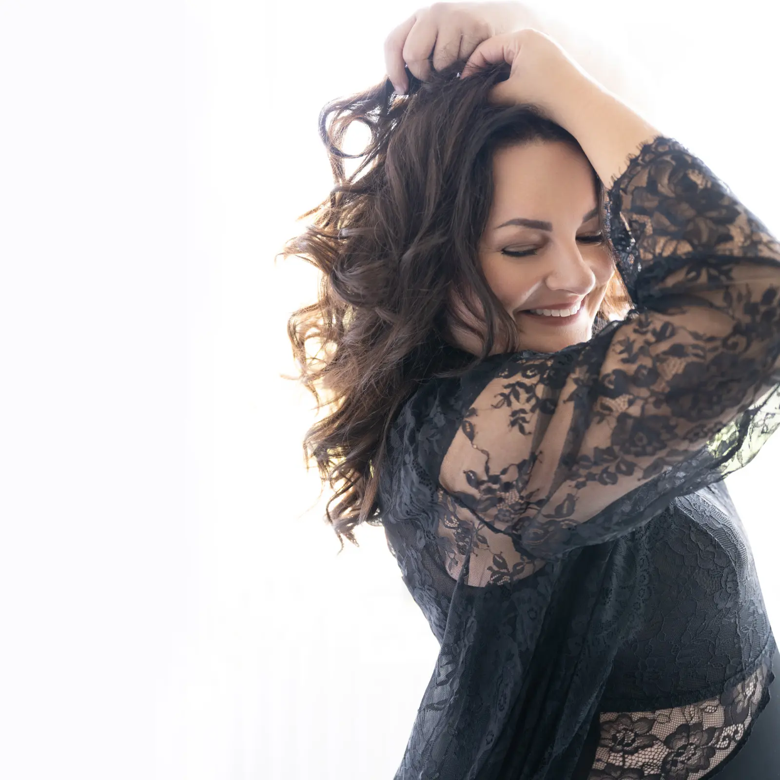 Smiling woman with long curly dark hair wearing a sheer black lace top, posing with one hand in her hair and eyes closed against a bright white background.