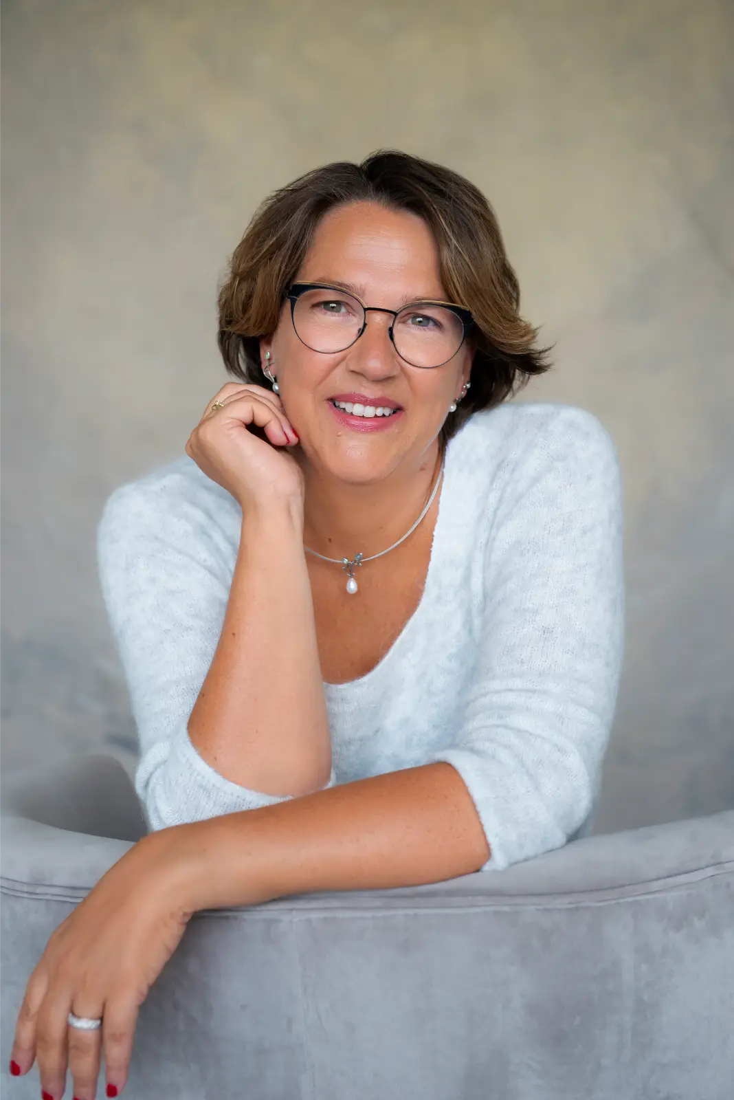 Smiling middle-aged woman with short brown hair and glasses wearing a white sweater, a pearl necklace with a pendant, earrings, and a ring, resting her arm on the back of a light grey chair against a neutral background.