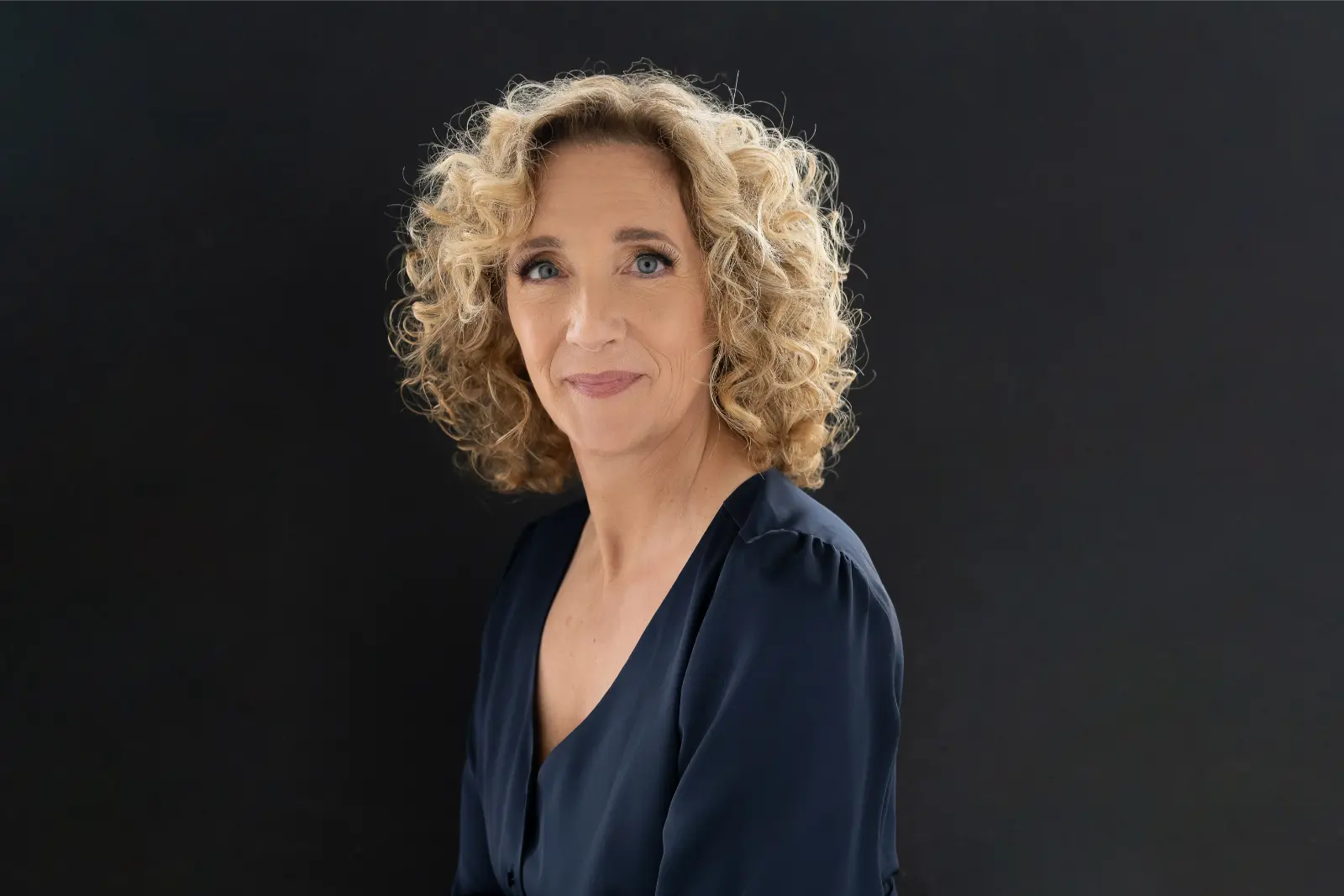 Portrait of a woman with curly blonde hair and blue eyes wearing a dark navy blouse, posing against a solid black background.