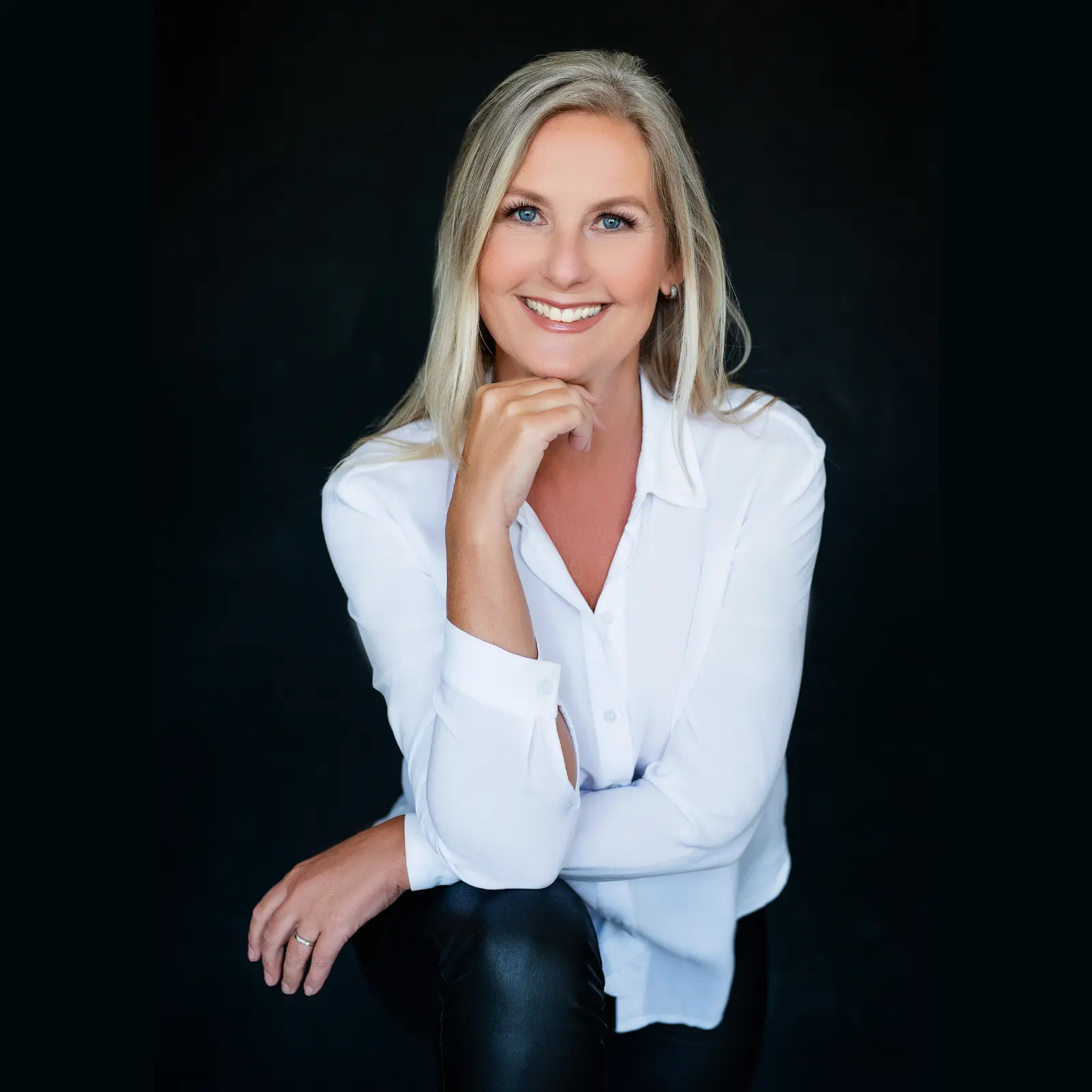 Smiling woman with long blond hair and blue eyes wearing a white button-up shirt and black pants, posing with one hand under her chin against a dark background.