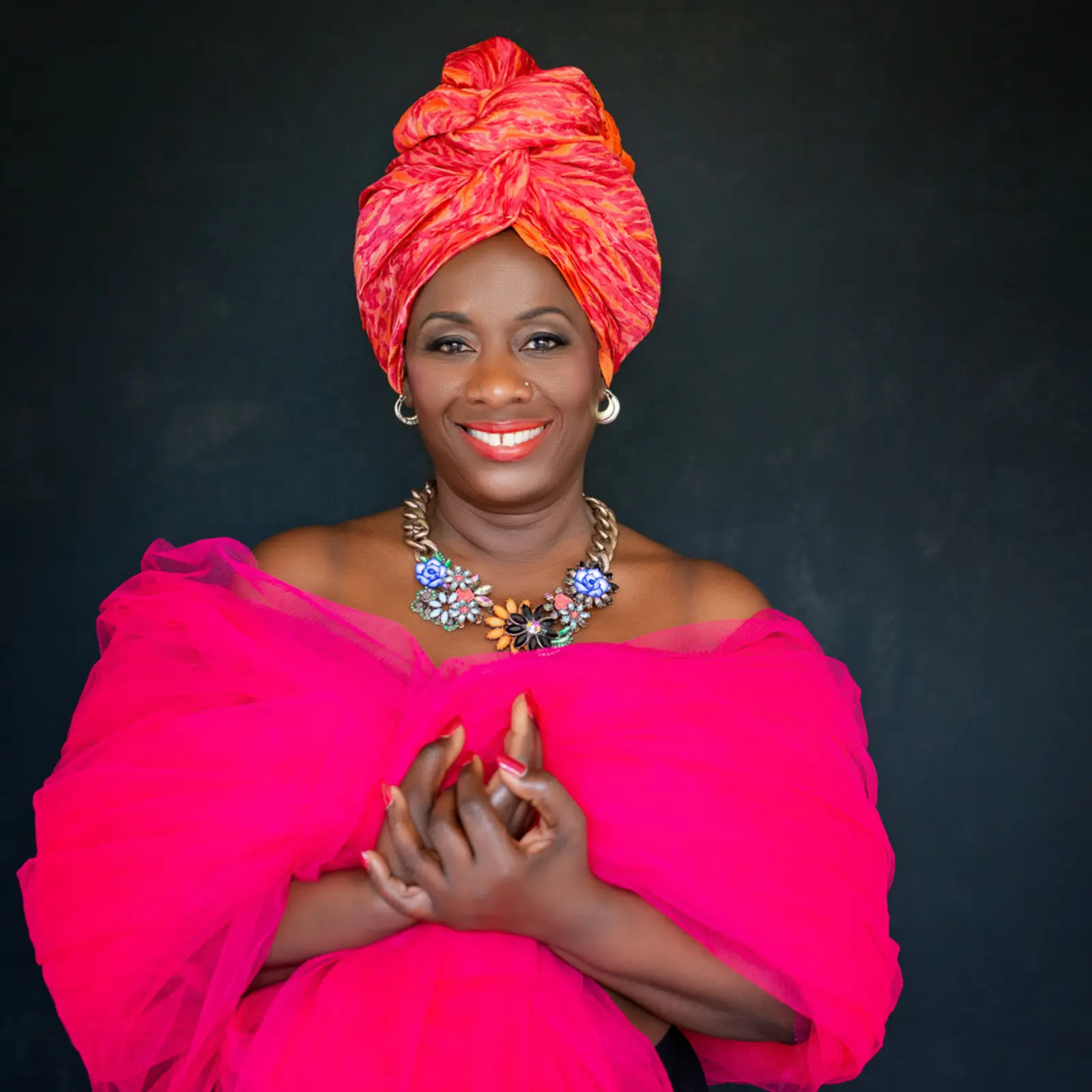 Smiling woman wearing a bright red and orange headwrap, a vibrant pink off-the-shoulder dress, and a colorful floral statement necklace, posed against a dark background.