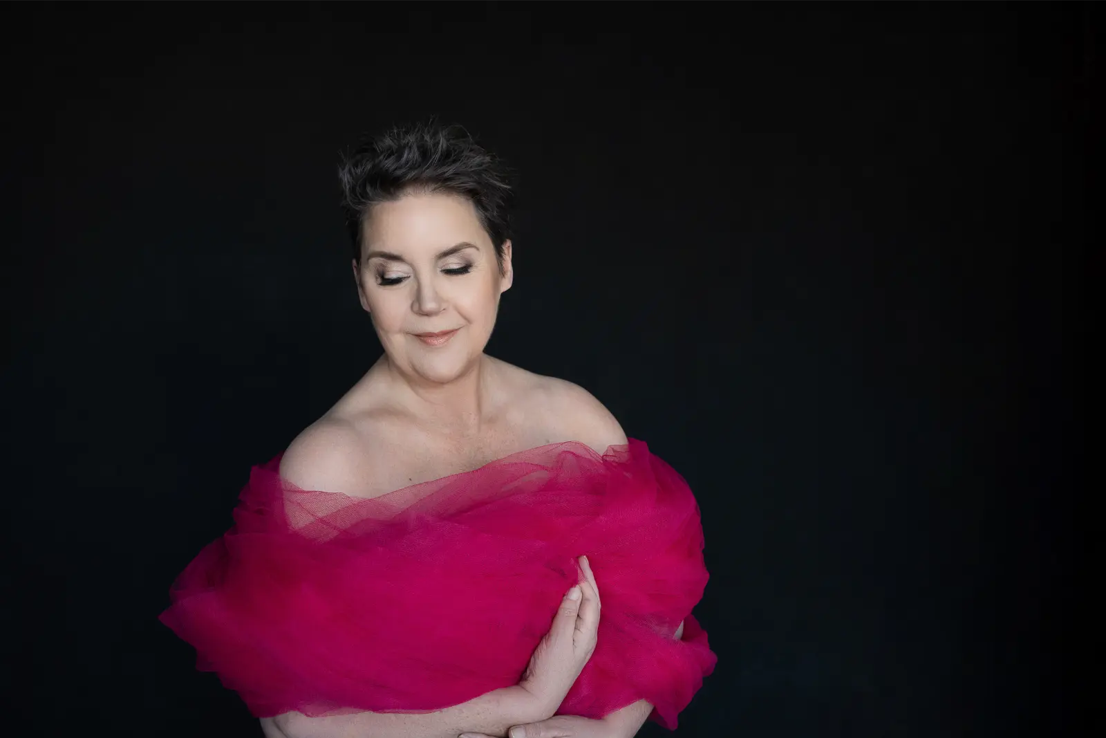 Woman with short dark hair and closed eyes, wrapped in a voluminous bright pink sheer fabric, against a plain black background.