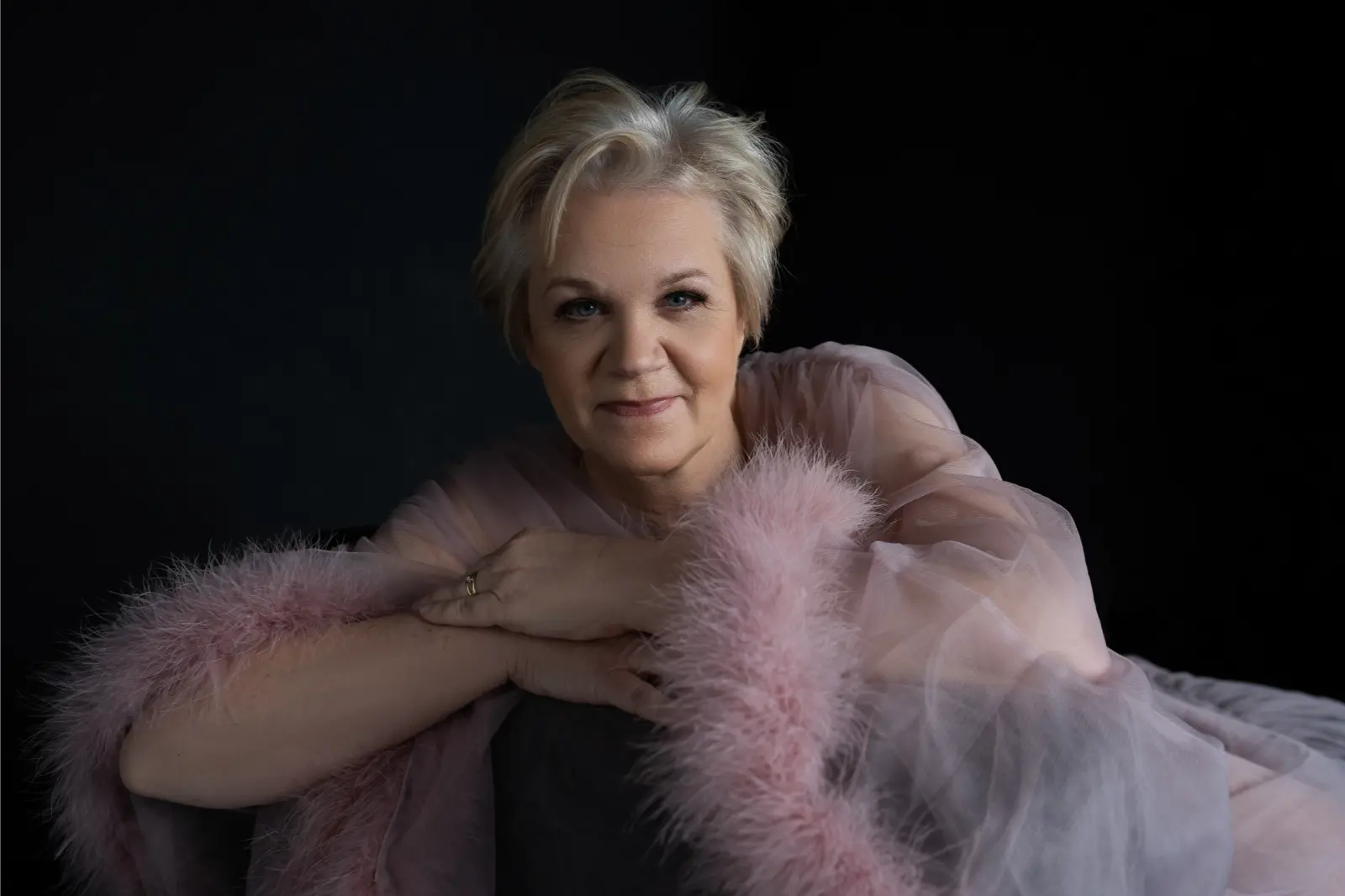 Portrait of a middle-aged woman with short blonde hair smiling softly, wearing a sheer pink garment with fluffy trim, against a dark background.