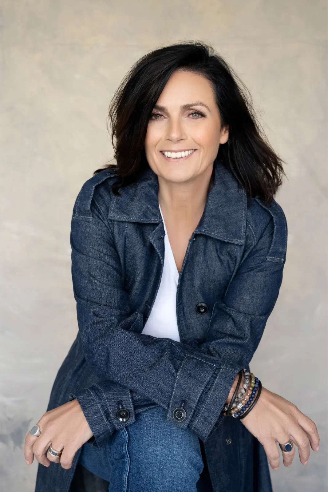 Smiling woman with shoulder-length dark hair wearing a dark denim jacket over a white top and blue jeans, sitting with arms crossed and wearing multiple rings and beaded bracelets on her wrists against a beige background.