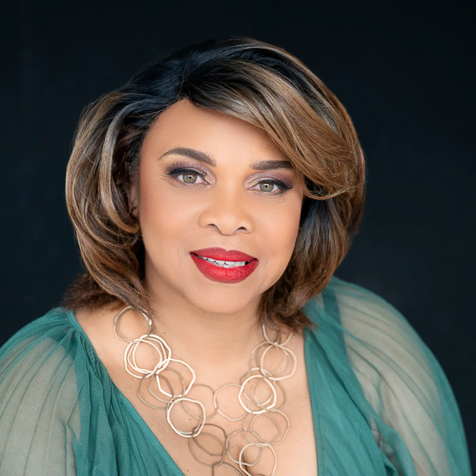 Portrait of a woman with short highlighted hair wearing red lipstick, a green sheer top, and a gold necklace with linked circles, against a dark background.