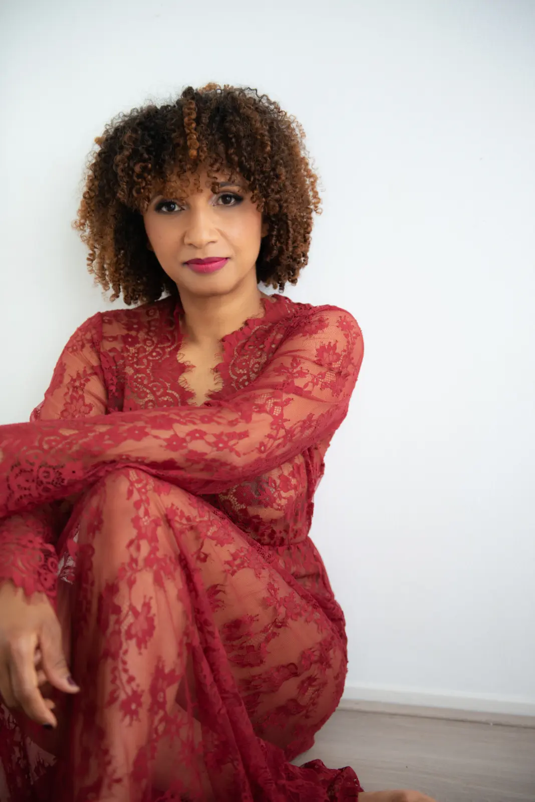 Woman with curly hair seated on the floor against a white wall, wearing a sheer red lace dress.