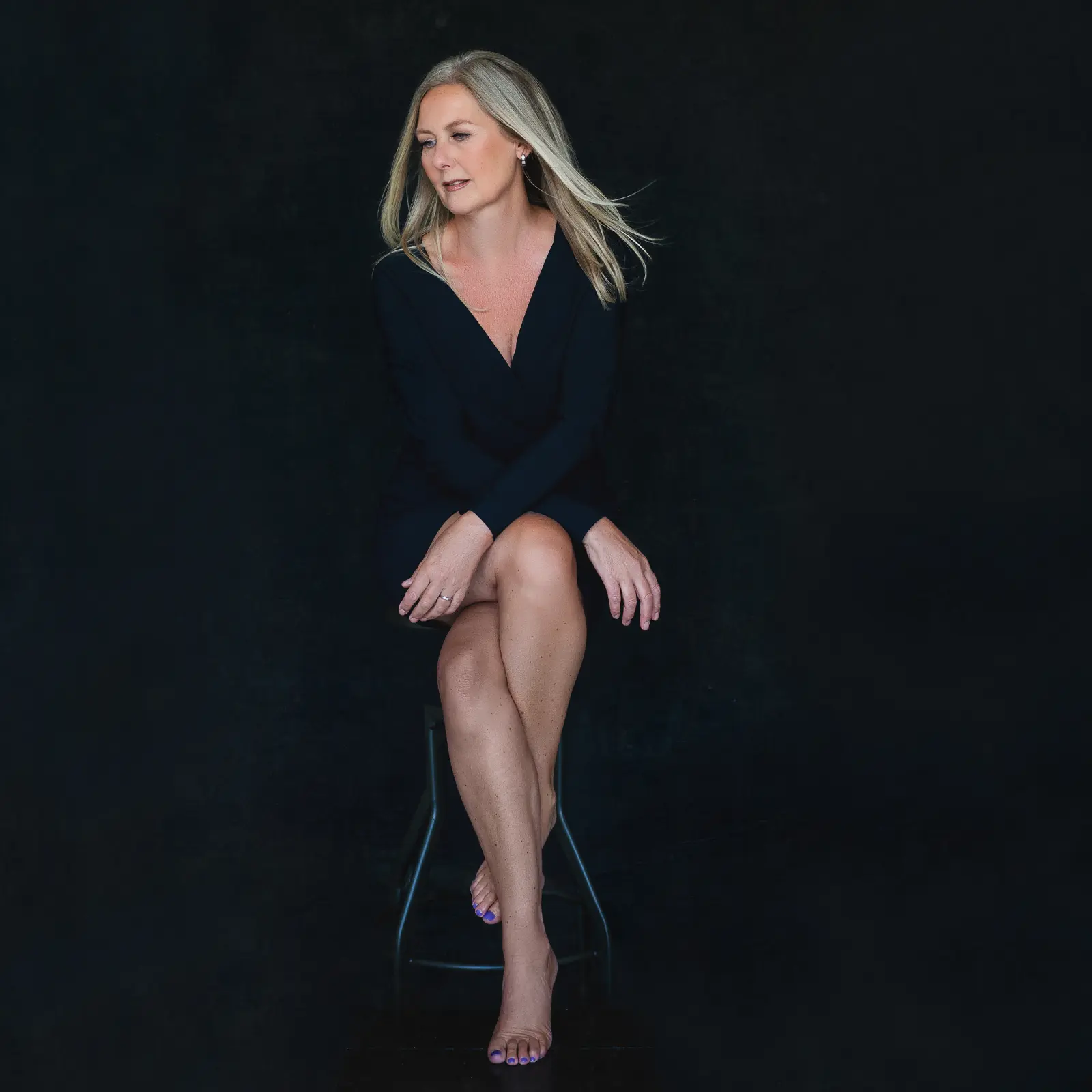 Blonde woman with shoulder-length hair wearing a black dress sitting barefoot on a black stool against a dark background, looking to her left with her hands resting on her crossed legs.