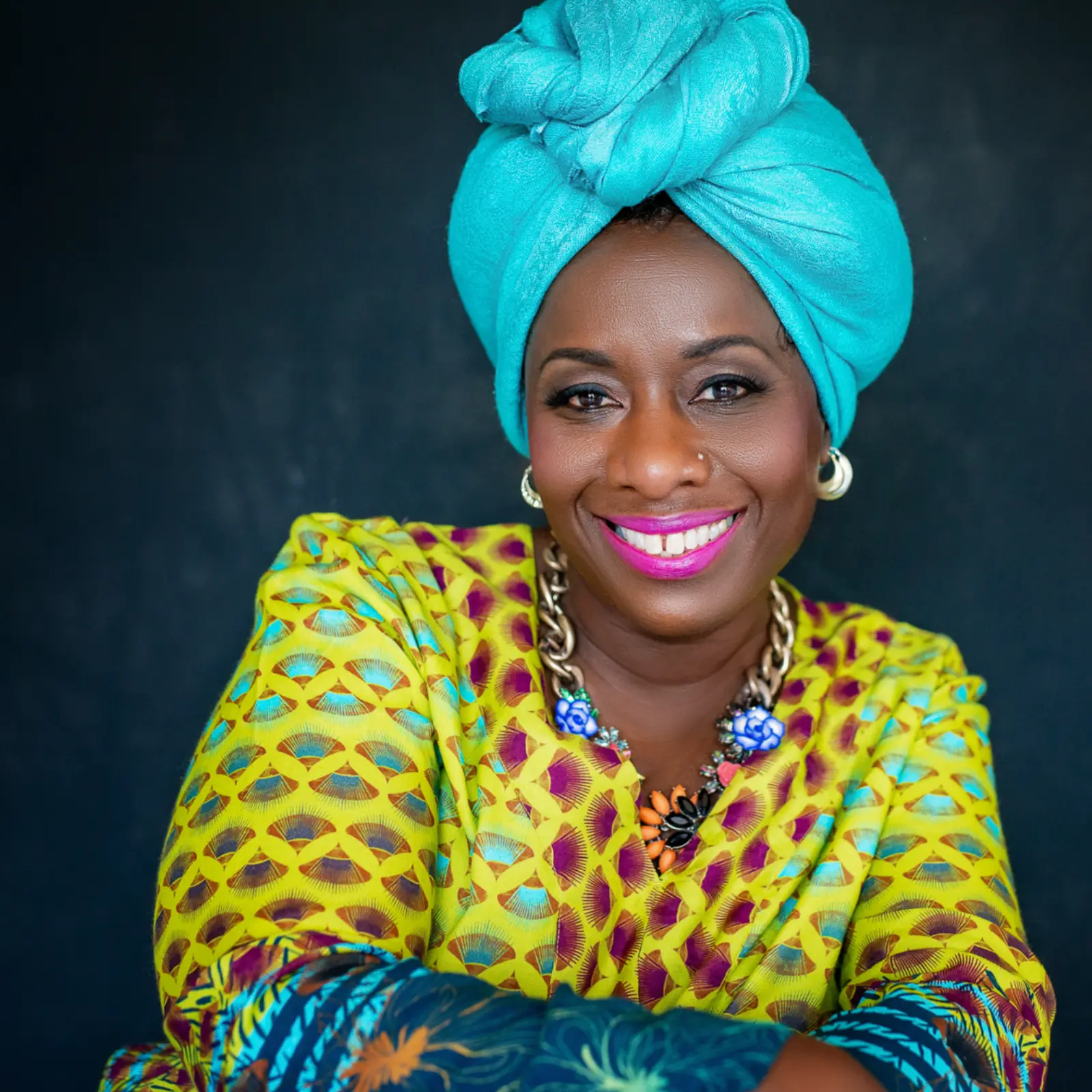 Smiling woman wearing a bright turquoise headwrap and colorful patterned clothing with a chunky flower necklace against a dark background.