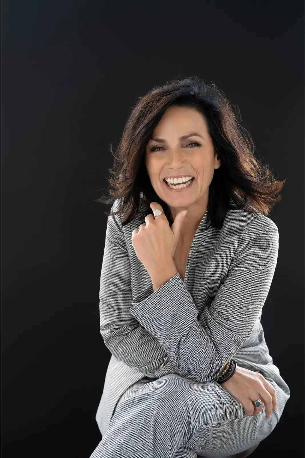 Smiling woman with shoulder-length dark hair wearing a gray checkered suit sitting against a black background, resting her chin on her hand.