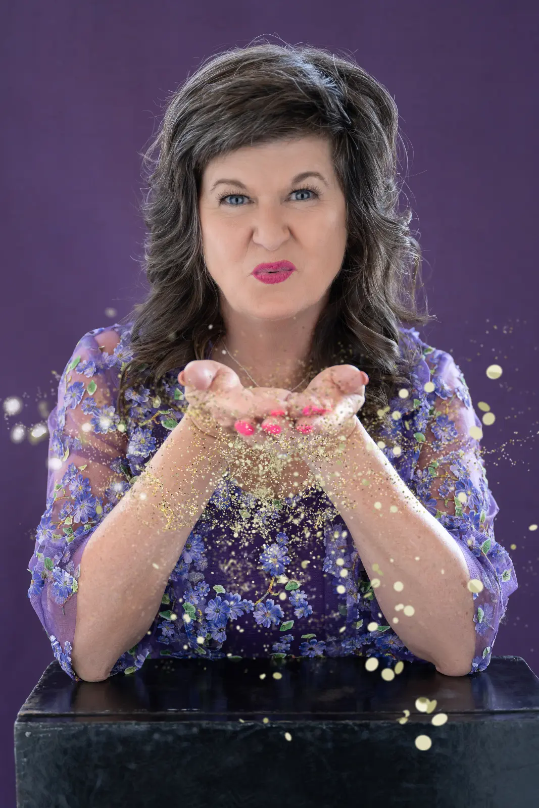 A woman with wavy dark hair and bright pink lipstick wearing a sheer purple blouse with floral embroidery blowing golden glitter from her cupped hands against a purple background.