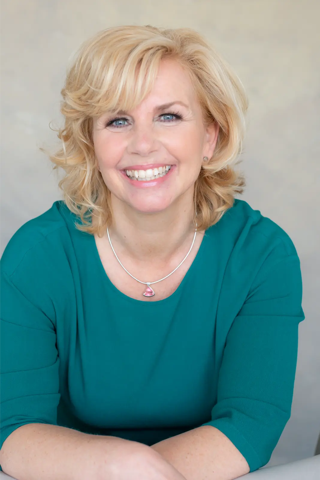 Smiling middle-aged woman with short blonde hair and blue eyes wearing a teal top and a silver necklace with a pink pendant, sitting with arms crossed on a table.