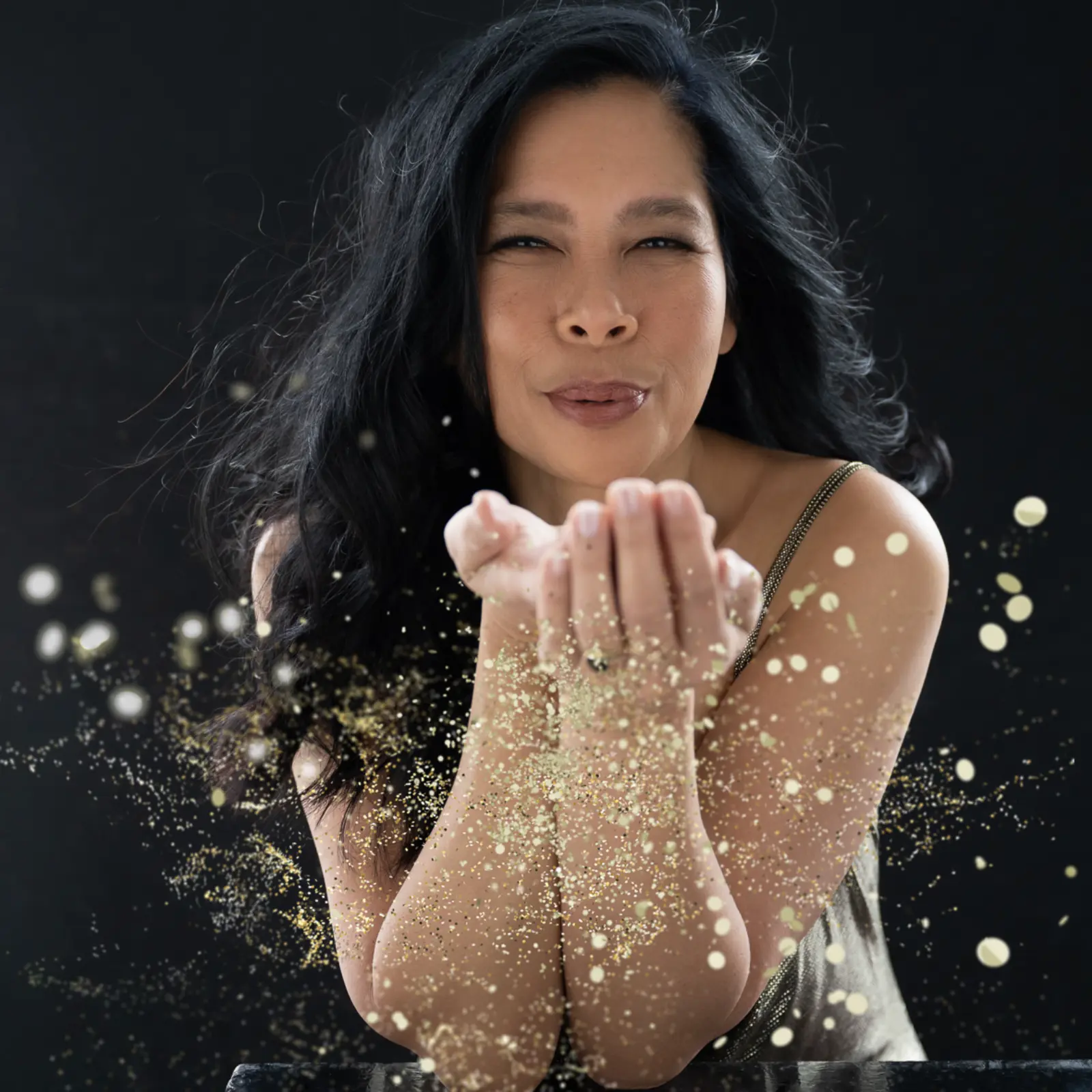 Middle-aged woman with long black hair blowing golden glitter from her cupped hands against a dark background.