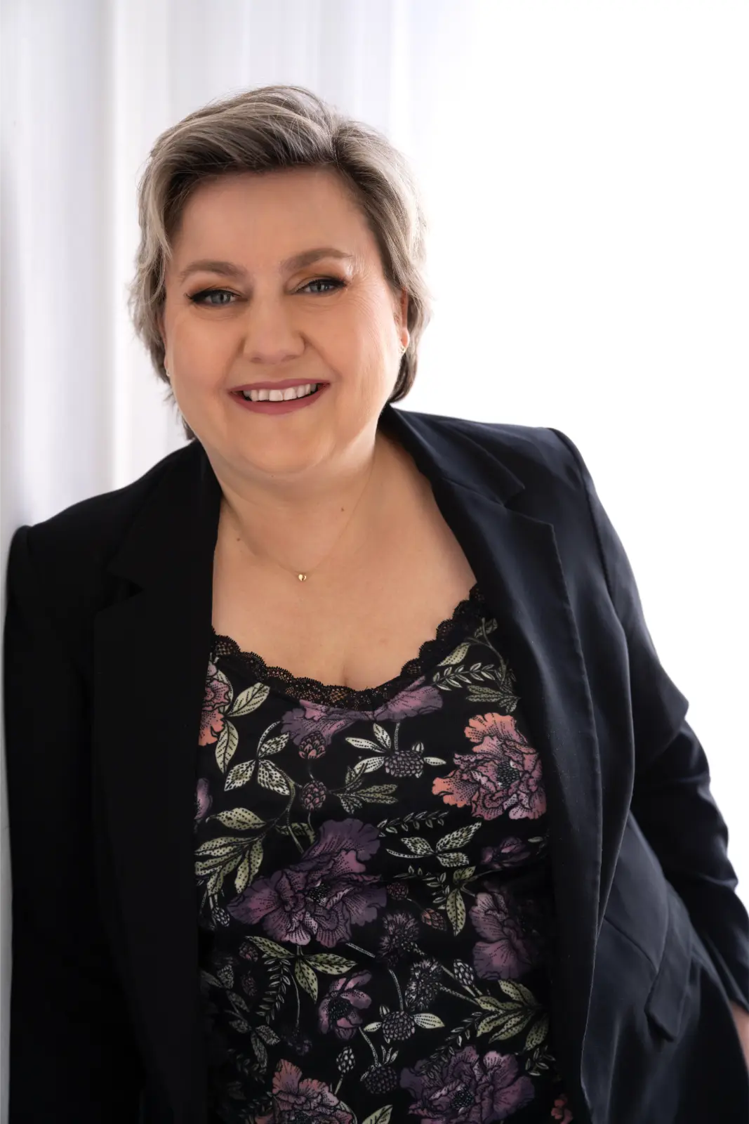 Smiling woman with short gray hair wearing a black blazer over a floral patterned top with purple and pink flowers, standing in front of a bright white background.